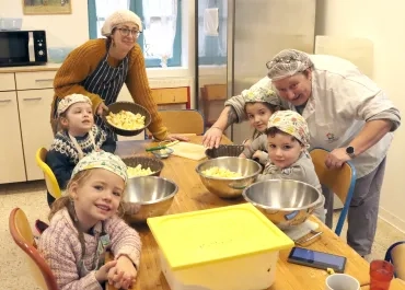 un groupe d'enfants et deux adultes avec des charlottes et des tabliers montrent des fruits en cuisine