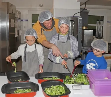 un animateur et trois enfants avec charlottes et tabliers mettent des légumes dans des plats en cuisine