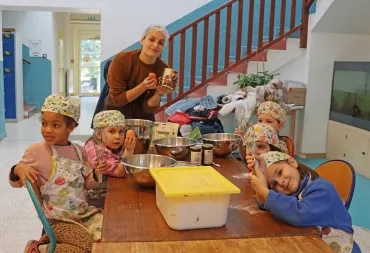 un groupe d'enfant et leur animatrice sont en cuisine avec des tabliers et des charlottes et montrent leurs ustensiles