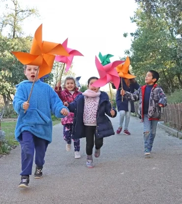 un goupe d'enfants et leur animatrice courent avec un grand moulin à vent en papier dans la main