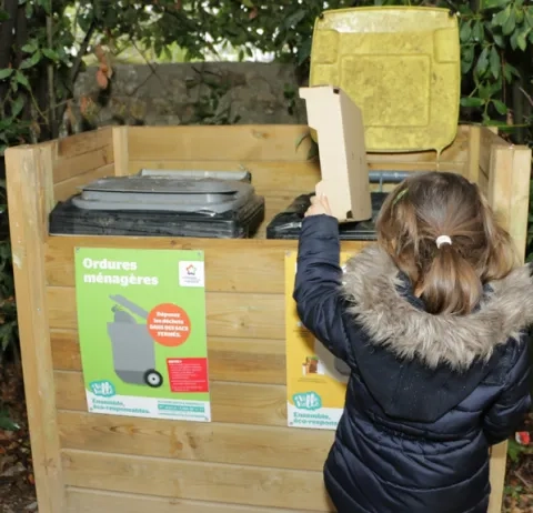 Une fillette jette un déchet dans un poubelle de tri placée dans un coffre de bois contenant cette poubelle de tri et une poubelle pour les déchets non recyclables