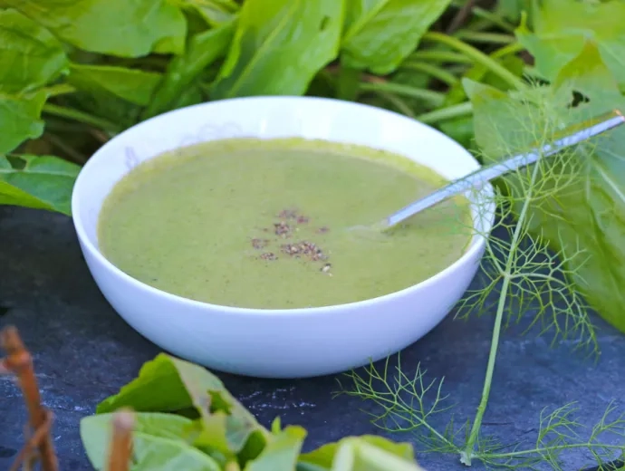 Une assiette creuse contenant de la soupe verte et une cuillere est installée sur une ardoise avec des légumes verts autour