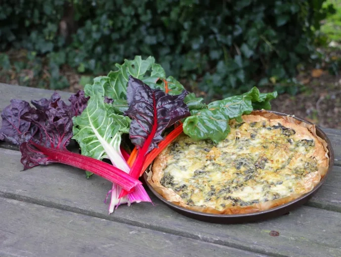 Une tarte aux blettes ainsi qu'un bouquet de blettes sont posés sur une table en bois