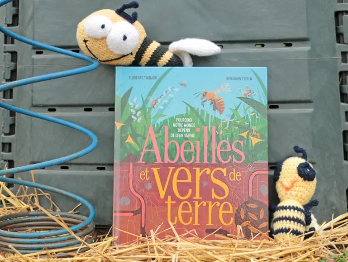 Le livre Abeilles et vers de terre est présenté face couverture, appuyé contre un composteur et entouré de deux peluches en forme d'abeilles