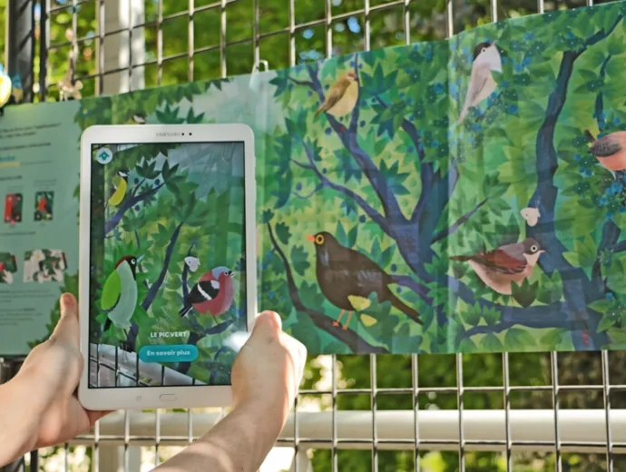 Une tablette numérique vise un des oiseaux présents sur le livre Ecoute les oiseaux déplié et exposé sur une grille