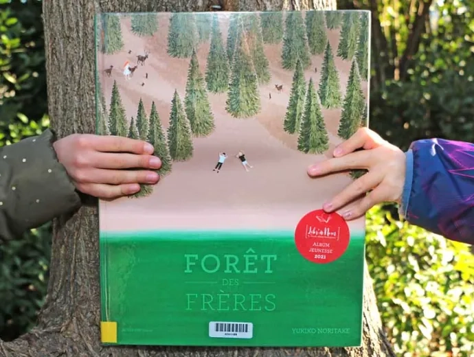 Le livre Forêt des frêres est présenté face couverture, tenu par deux mains appartenants à deux enfants différents, chacun d'eux le tenant d'un côté du tronc d'un arbre