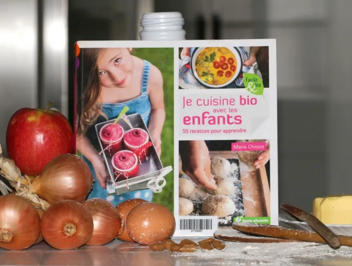 Le livre Je cuisine bio avec les enfants est exposé dans une cuisine et entouré d'aliments frais comme du beurre, des pommes, des oignons