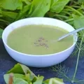 Une assiette creuse contenant de la soupe verte et une cuillere est installée sur une ardoise avec des légumes verts autour