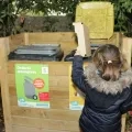 Une fillette jette un déchet dans un poubelle de tri placée dans un coffre de bois contenant cette poubelle de tri et une poubelle pour les déchets non recyclables