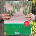 Le livre Forêt des frêres est présenté face couverture, tenu par deux mains appartenants à deux enfants différents, chacun d'eux le tenant d'un côté du tronc d'un arbre