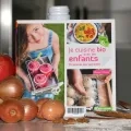 Le livre Je cuisine bio avec les enfants est exposé dans une cuisine et entouré d'aliments frais comme du beurre, des pommes, des oignons