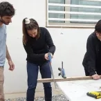 Une adulte utilise une scie à bois pour couper un tasseau maintenu par une autre adulte. Un animateur supervise.