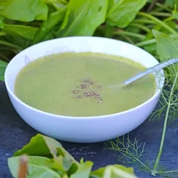 Une assiette creuse contenant de la soupe verte et une cuillere est installée sur une ardoise avec des légumes verts autour