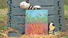 Un livre est posé devant un composteur avec deux abeilles en tricot posées dessus et à côté ainsi qu'un outil permettant d'aérer le compost