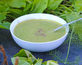 Une assiette creuse contenant de la soupe verte et une cuillere est installée sur une ardoise avec des légumes verts autour