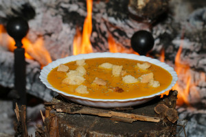 une assiette creuse avec une soupe orangée et des croutons de pain est présentée devant un feu de cheminée allumé