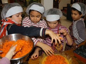 Des enfants équipés de charlottes mélangent des carottes rapées avec d'autres ingrédients pour préparer des cakes aux carottes